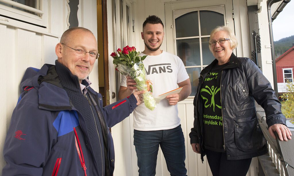 Amer Hidic ( i midten) fikk blomster og hovedpremie i Verdensdagslotteriet i Hemnes, overrakt på døren. De som banket på var leder i Mental Helse Hemnes, Birgit Fjelldal og sekretær Øyvind Arntsen.