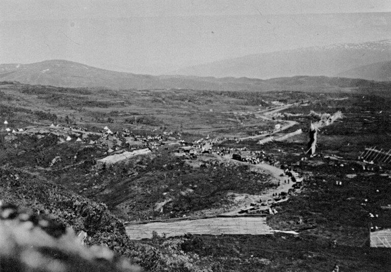 Bildet viser en sammenkomst på fjellet med mange biler og mye folk. Den nye vegen er i bruk, og snøskjermer er satt opp. Skjæringa ved dagens Fjellstue er under arbeid.I bakgunnen skimtes brøytestasjonen på nordsida. Bildet er tatt fra Svalvasshåjen og nordover.